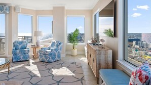 A spacious living room with a large window offering a city view, two patterned armchairs, a round coffee table, and a wooden cabinet. - Four Bedroom Luxury Condo with Spectacular Ocean & Bay Views (Ocean City)