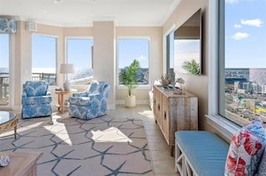 A spacious living room with a large window offering a city view, two patterned armchairs, a round coffee table, and a wooden cabinet.