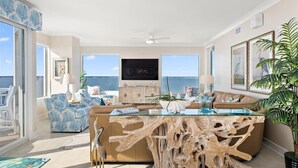 A spacious living room with a large glass coffee table, a beige sofa, and a view of the ocean. - Four Bedroom Luxury Condo with Spectacular Ocean & Bay Views (Ocean City)