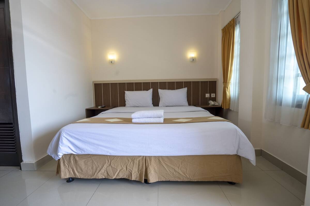 Una cama bien hecha con colcha blanca y color beige, dos mesitas de noche con lámparas, y una ventana con cortinas.
