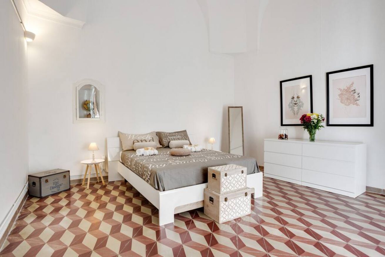A bedroom with a checkered floor, a bed with multiple pillows, a bedside table with a lamp, a dresser, and two framed pictures on the wall.