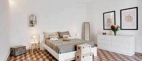 A bedroom with a checkered floor, a bed with multiple pillows, a bedside table with a lamp, a dresser, and two framed pictures on the wall.
