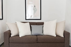 A brown sofa with four pillows, a framed artwork on the wall, and a plain white wall.