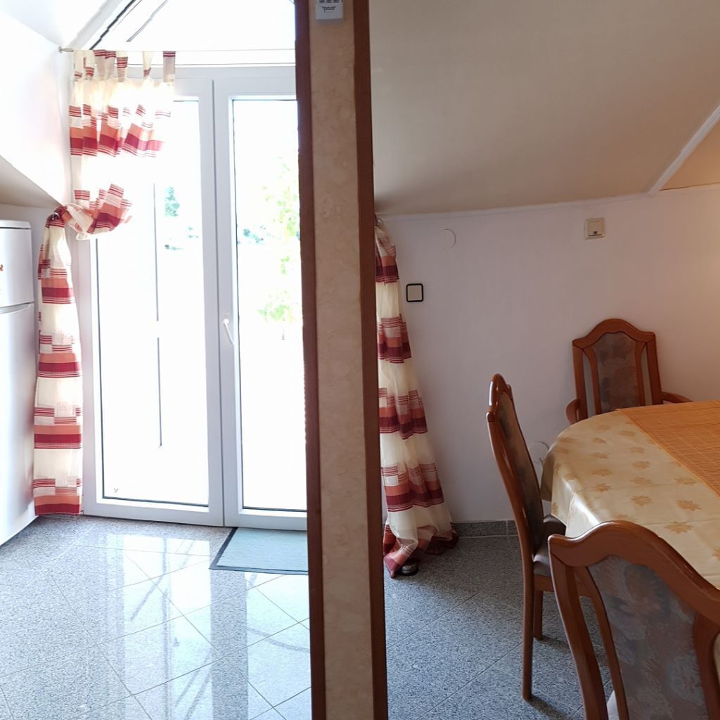 A room with a bed, a wooden chair, a table, a door with curtains, and a refrigerator.