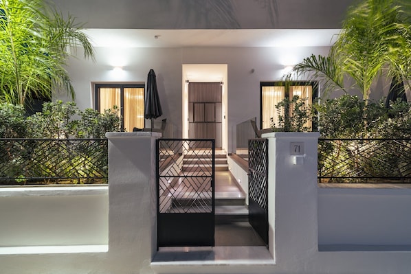 A modern building entrance with a staircase leading to a door, surrounded by greenery and a metal fence.