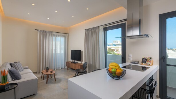 Een moderne keuken met een centraal eiland, een zithoek met een bank en een kleine tafel, en een woongedeelte met een televisie en een plant.