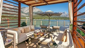 A covered outdoor seating area with a sofa, chairs, a table with food and drinks, and a view of a lake and mountains. - NG Phaselis Bay - All inclusive (Kemer)