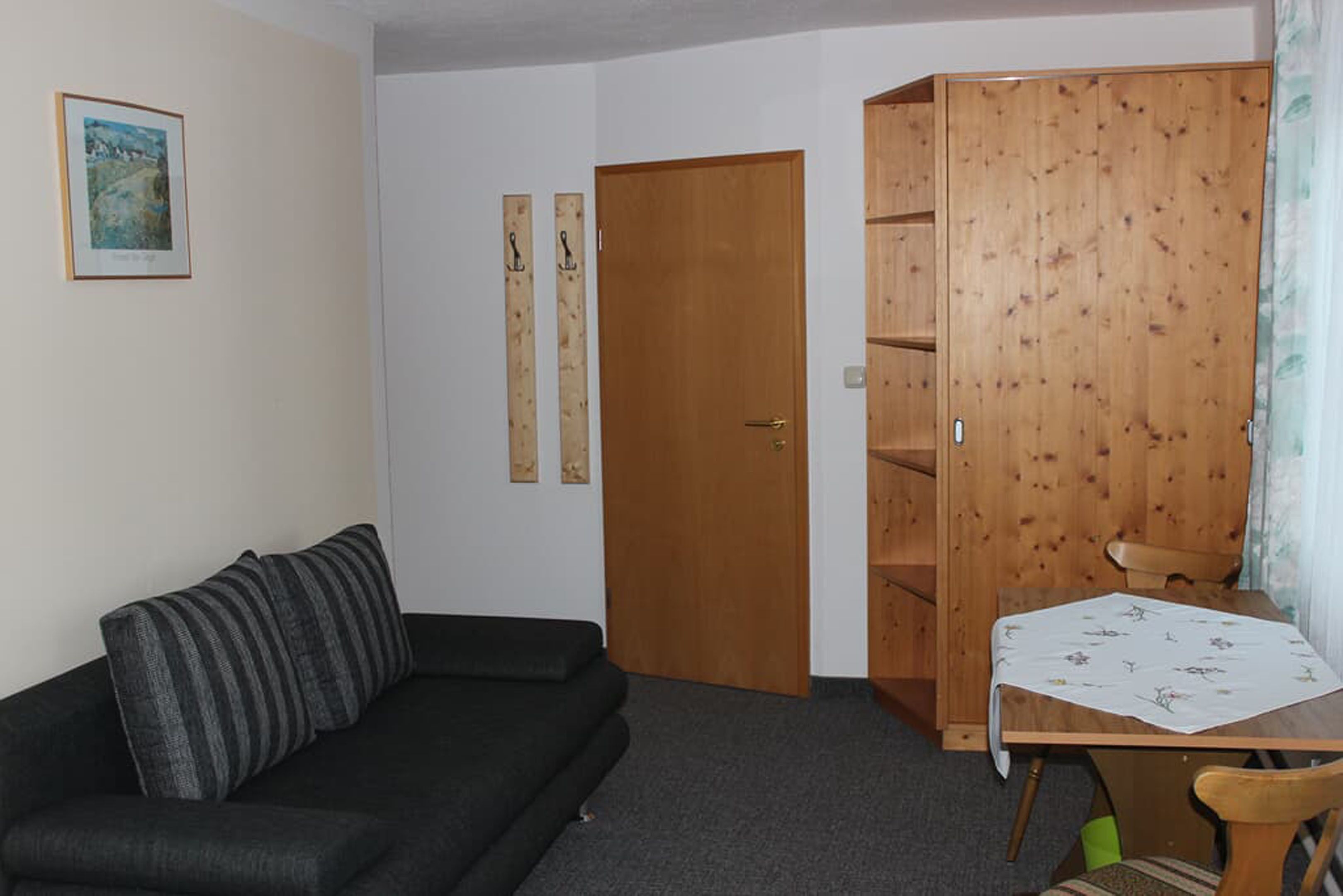 A room with a black sofa, a wooden wardrobe, a small table with a floral tablecloth, and a door.