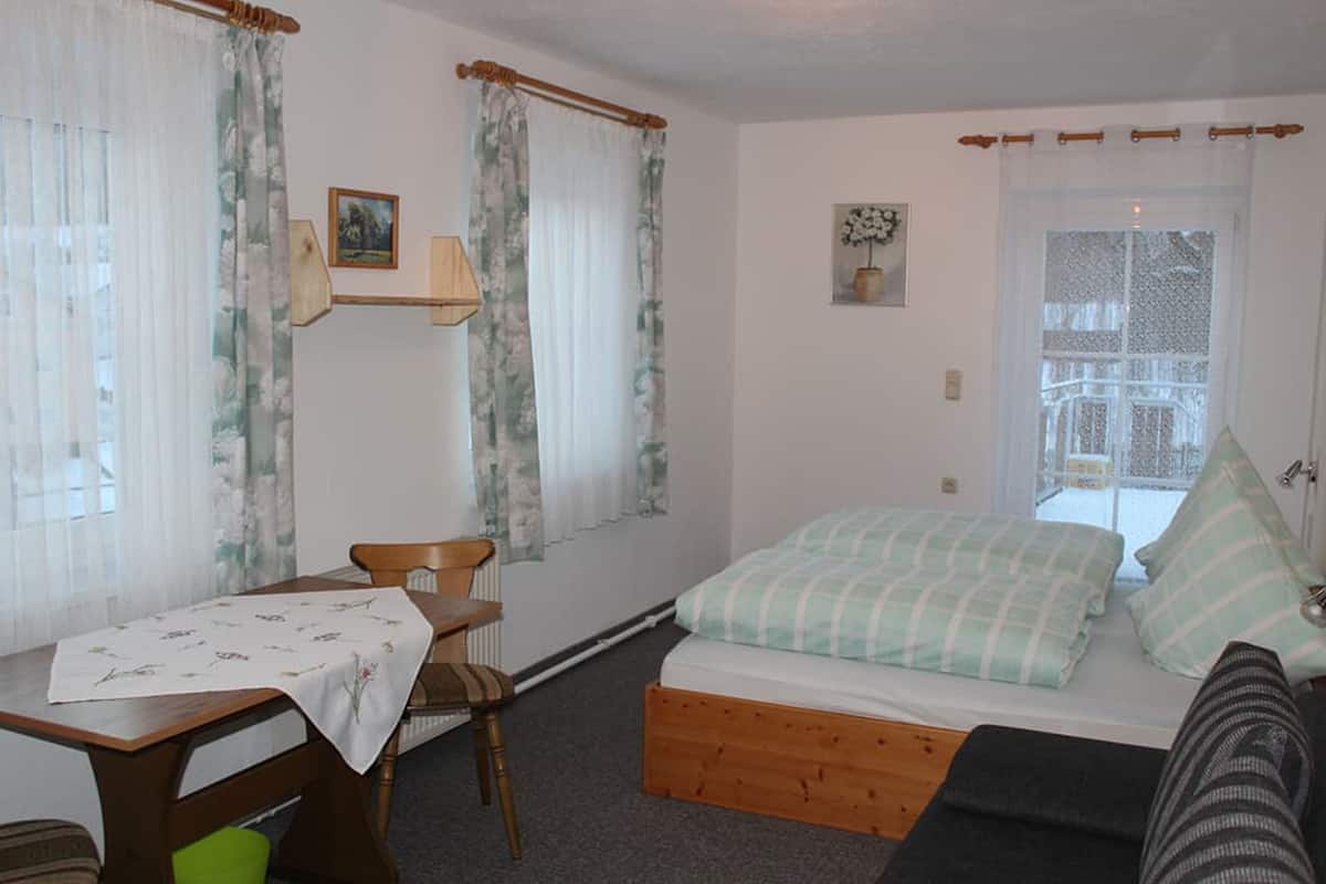 A small, cozy room with a bed, a wooden table, and a chair. There is a window with curtains and a view of the outside.