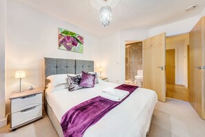A modern bedroom with a large bed, a nightstand, a bedside lamp, a chandelier, and a wooden door. - Paladin Place (Redhill)