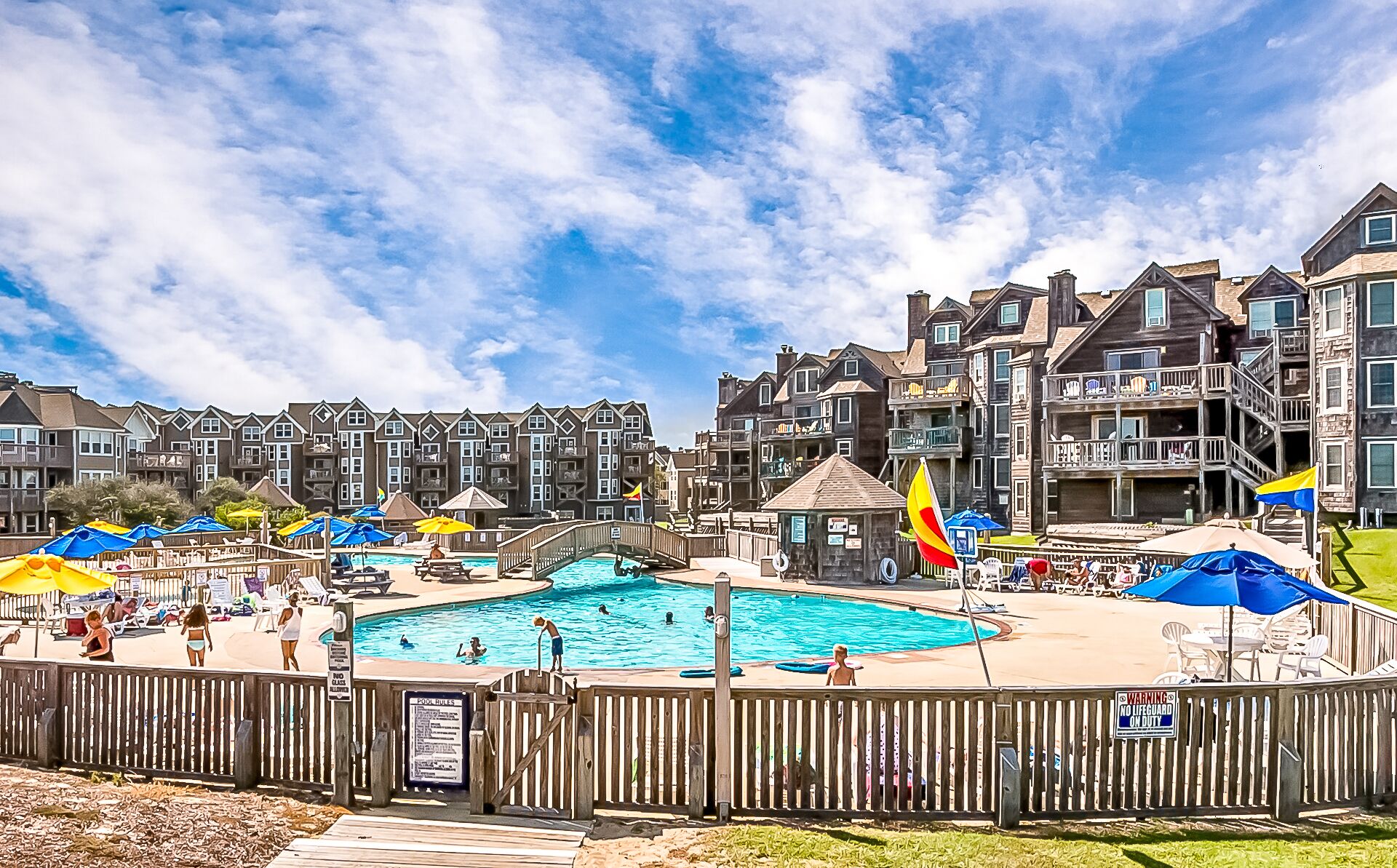 Un espace piscine agrémenté de plusieurs parasols, d’une terrasse en bois et offrant une vue sur des immeubles d’habitation en arrière-plan.