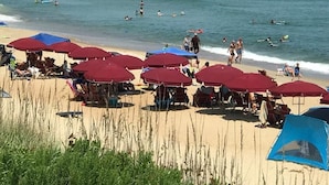 Uma praia com guarda-sóis vermelhos, um píer ao fundo e pessoas nadando e tomando sol.