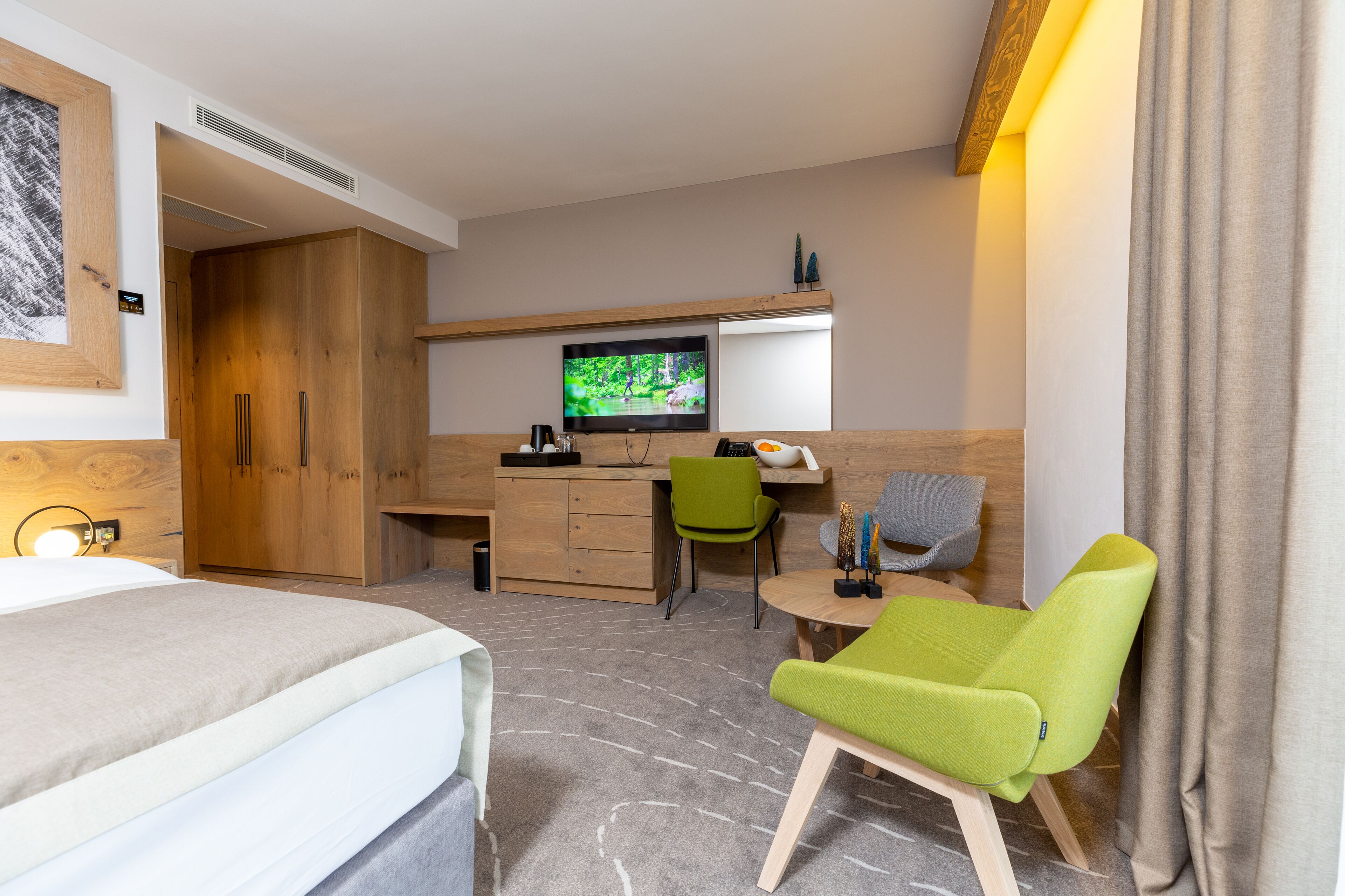 A hotel room with a bed, a desk with a television, a green chair, and a small table.