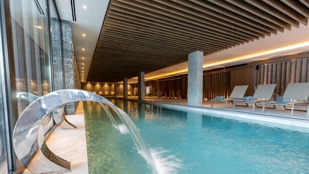 An indoor swimming pool with a waterfall feature, lounge chairs, and a modern architectural design. - Hotel Monti Spa&Wellness (Hadzici)