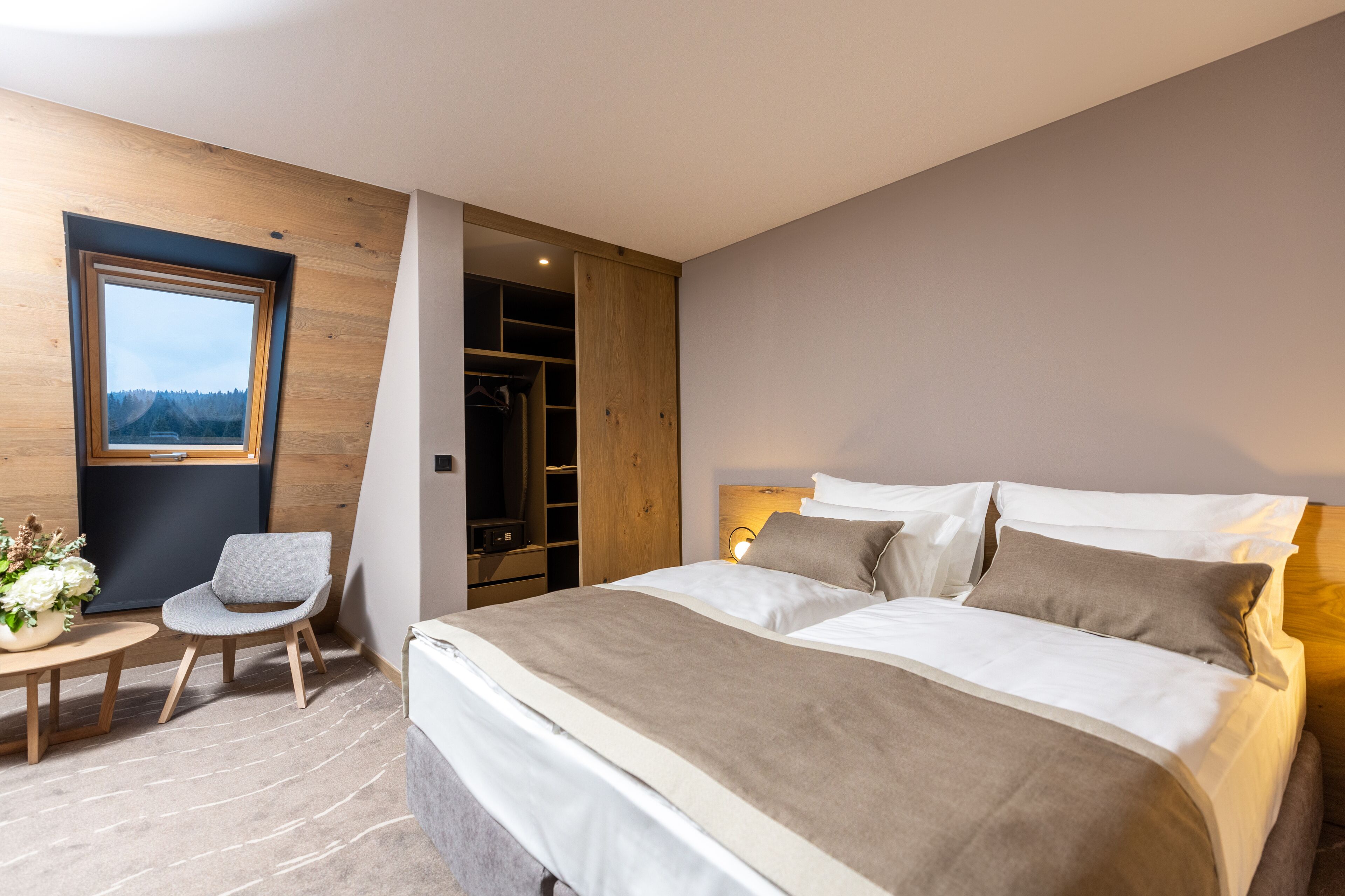 A modern bedroom with a large bed, a small seating area, and a closet with a view of the outdoors.