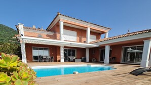Una casa de dos pisos con piscina, terraza de madera y mobiliario exterior.