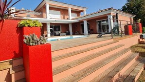 Una casa de dos pisos con piscina, macetas rojas y una escalera de madera.