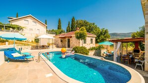 A swimming pool with lounge chairs and umbrellas, a stone building, and a view of the hills. - Dubrovnik Chalets - 2 Charming Stone Houses (Konavle)