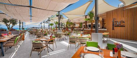 An outdoor dining area with tables and chairs, umbrellas, and a view of the ocean.