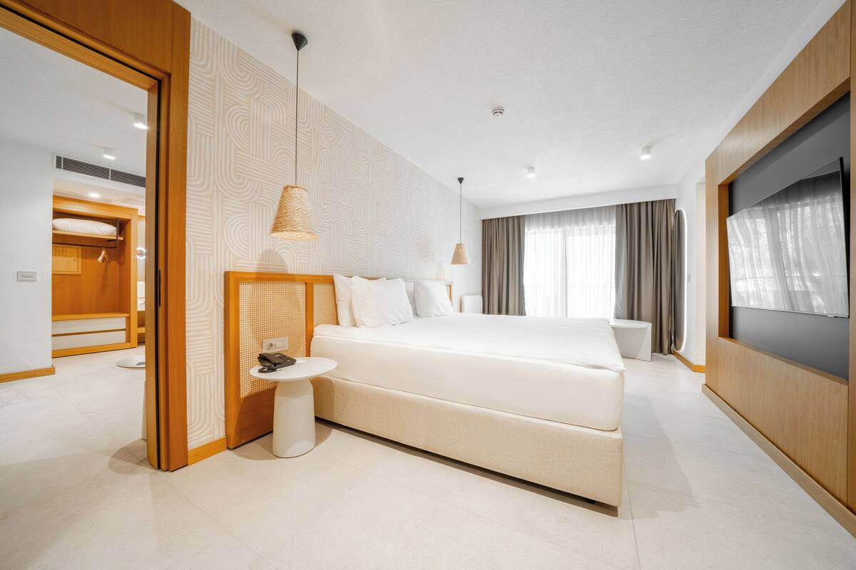 Quarto de hotel moderno com uma cama grande, mesa de cabeceira, televisão e banheiro visível através de uma porta aberta.
