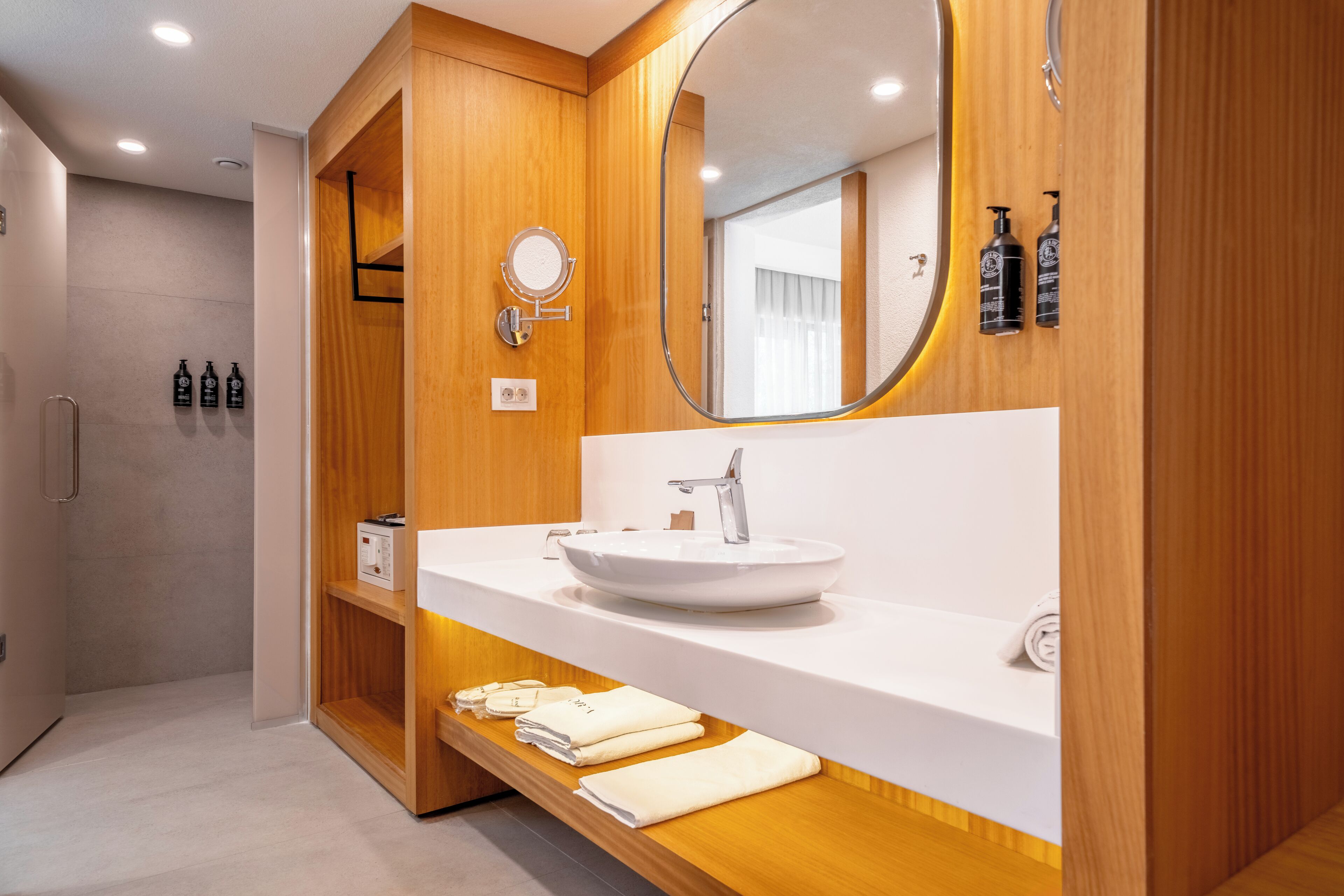 Een moderne badkamer met een houten wastafel, een ronde spiegel, een witte wasbak en netjes opgevouwen handdoeken op een plank.