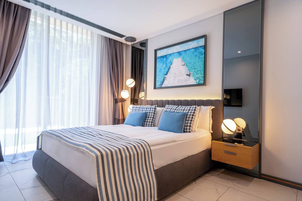 Quarto de hotel moderno com uma cama grande, mesa de cabeceira com abajur e um quadro na parede.