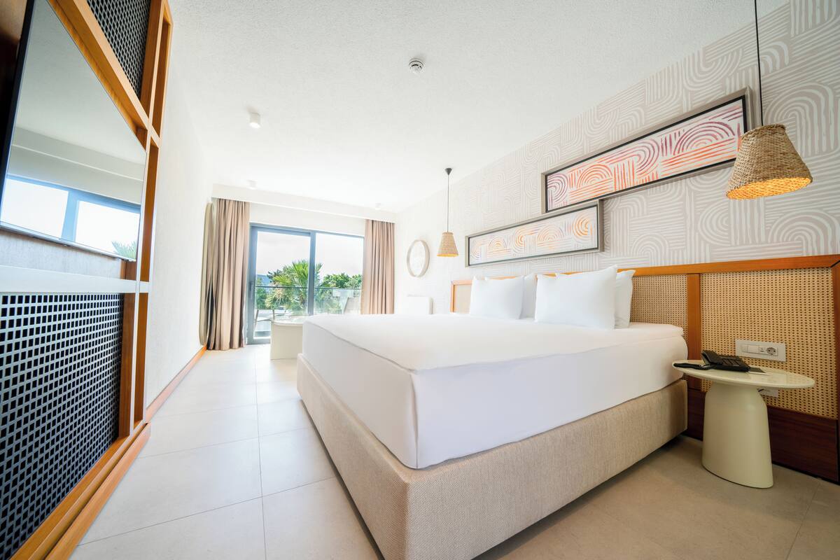 Quarto de hotel moderno com uma cama grande, uma mesinha com um telefone e vista para o exterior pela janela.