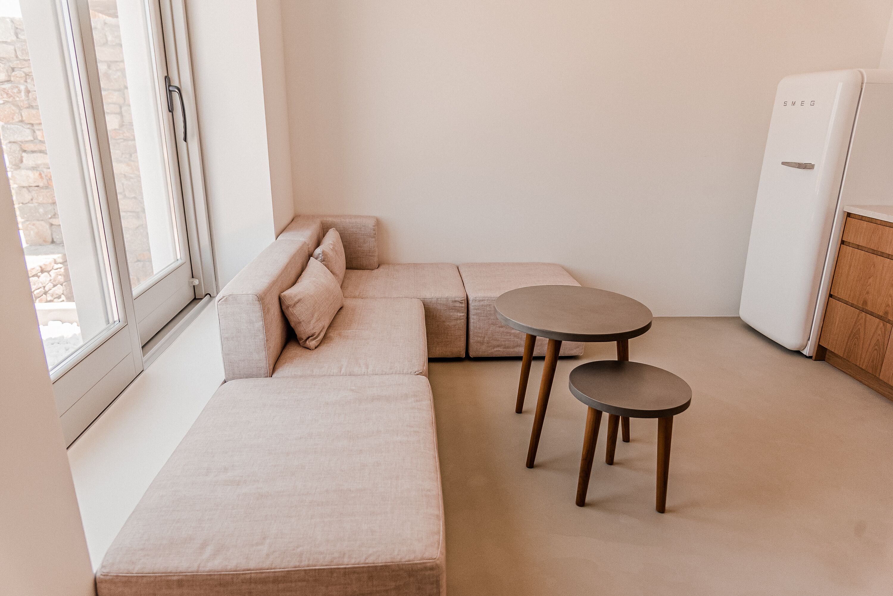 Une pièce moderne et minimaliste, avec un canapé d’angle beige, deux tables rondes et un réfrigérateur blanc.