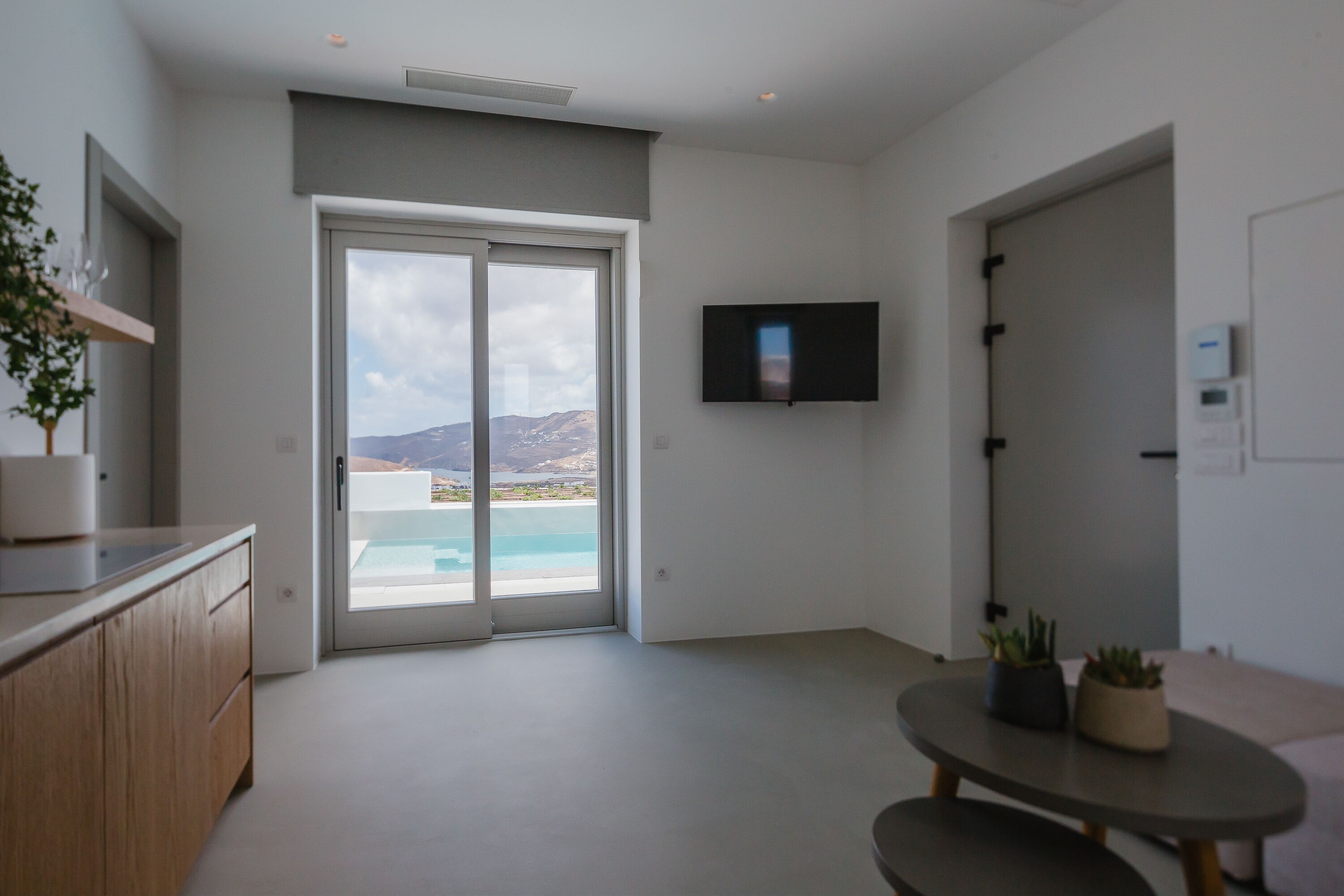 Un salon moderne doté d’une grande porte-fenêtre coulissante donnant sur un balcon avec vue sur une plage et des montagnes. Un téléviseur à écran plat est fixé au mur, et une petite table ronde est agrémentée de plantes en pot.
