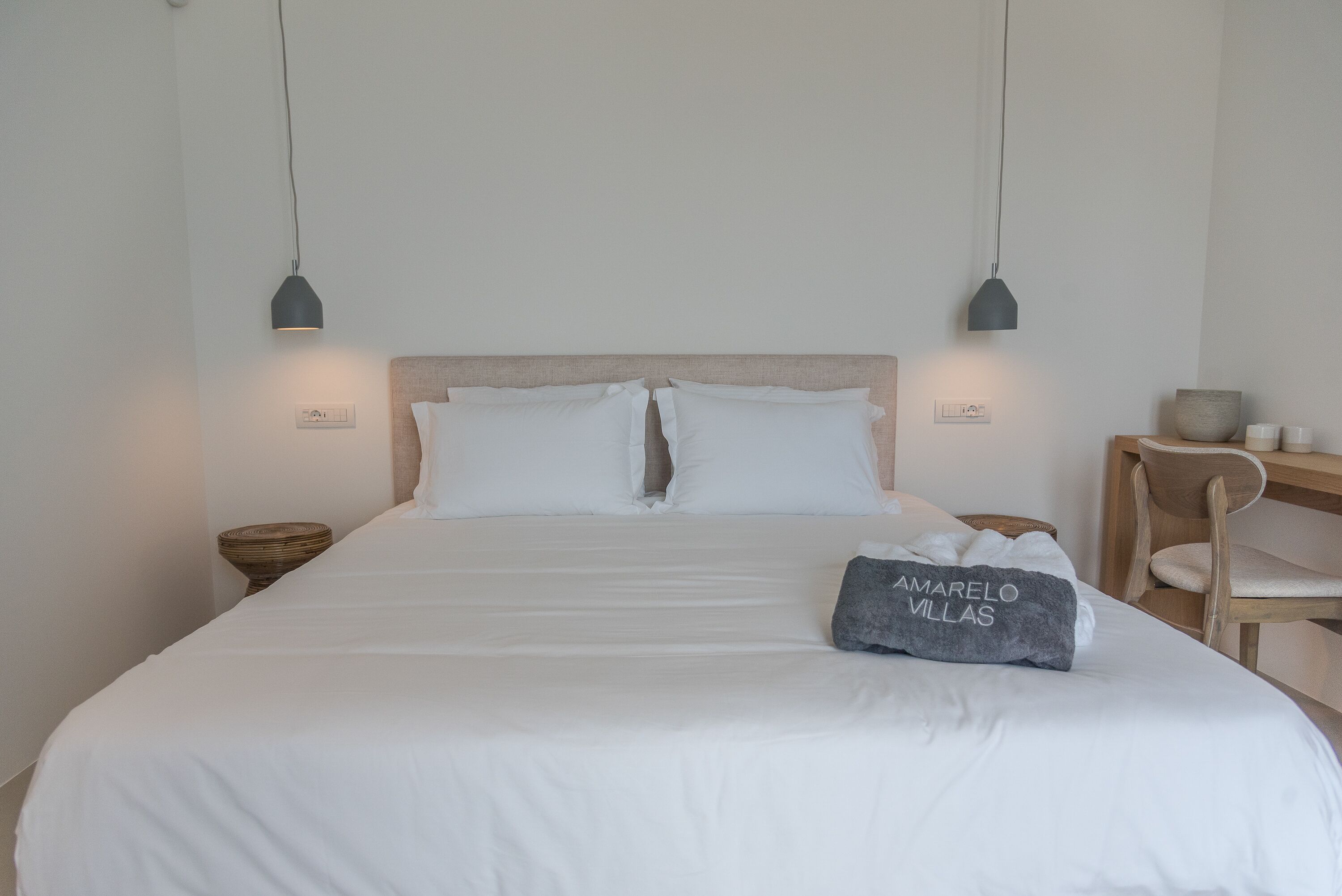 A neatly made bed with white linens and pillows, a grey pillow with "AMARELO VILLAS" text, two bedside tables with lamps, and a wooden chair.