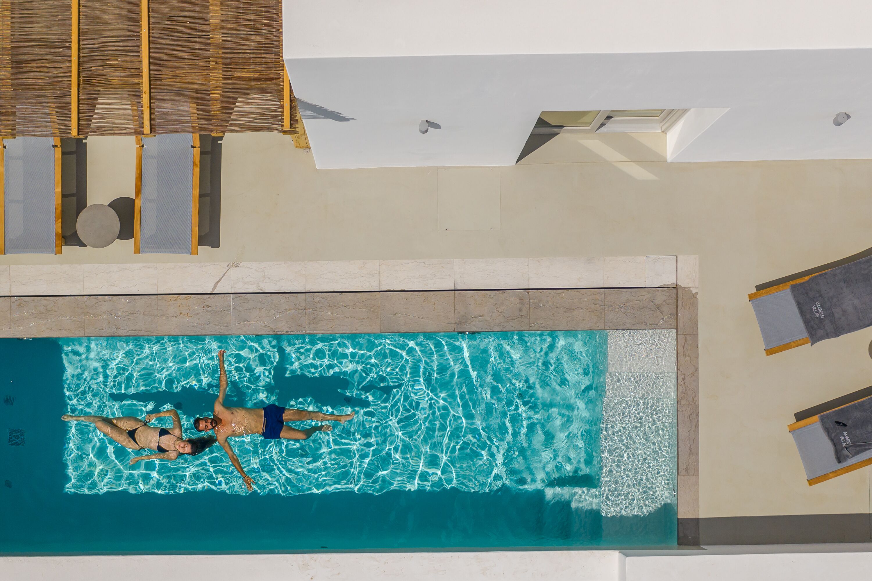 An aerial view of a swimming pool with two people floating, surrounded by sun loungers and a covered area.