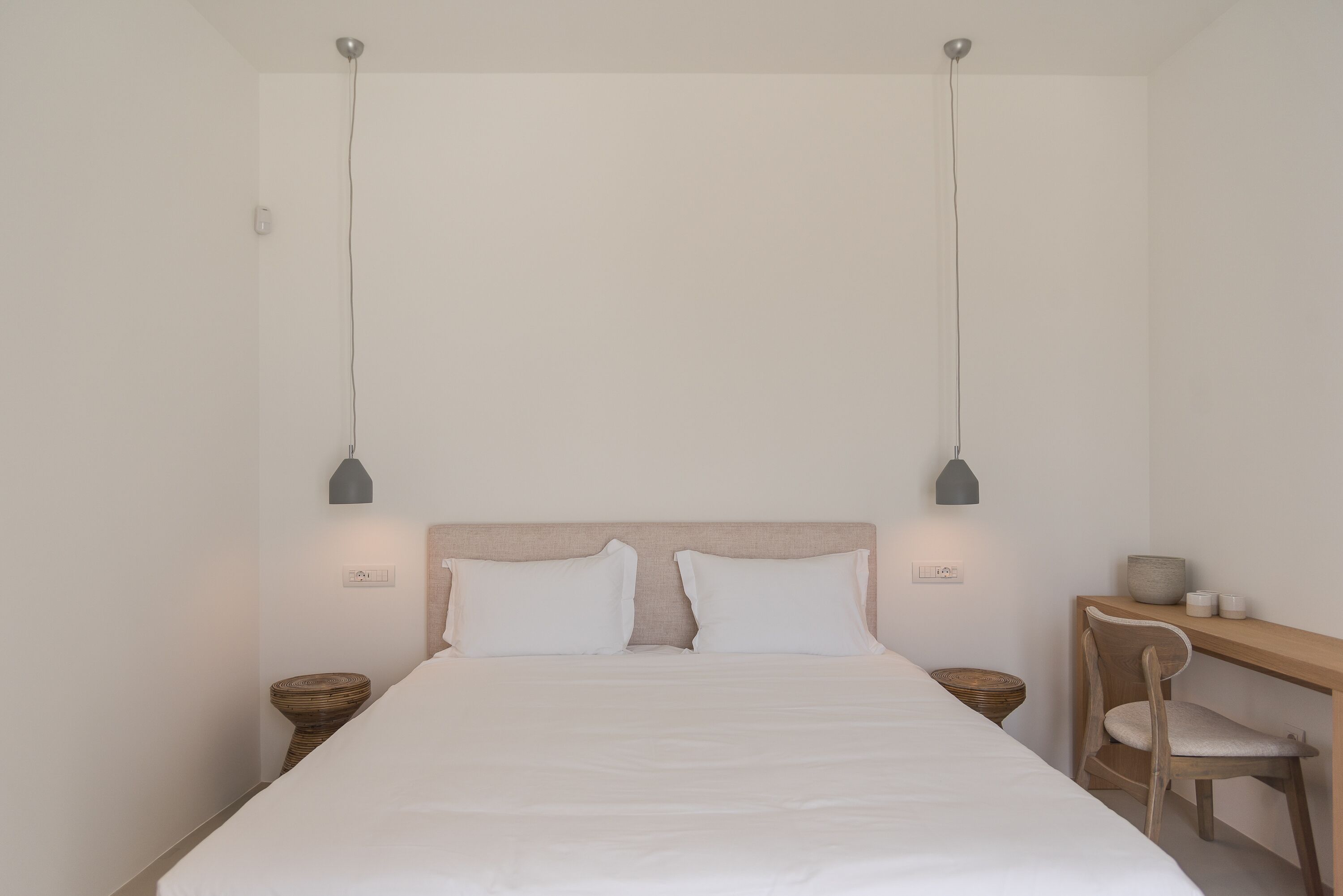 A minimalist bedroom with a bed, two bedside tables, a desk with a chair, and pendant lighting.