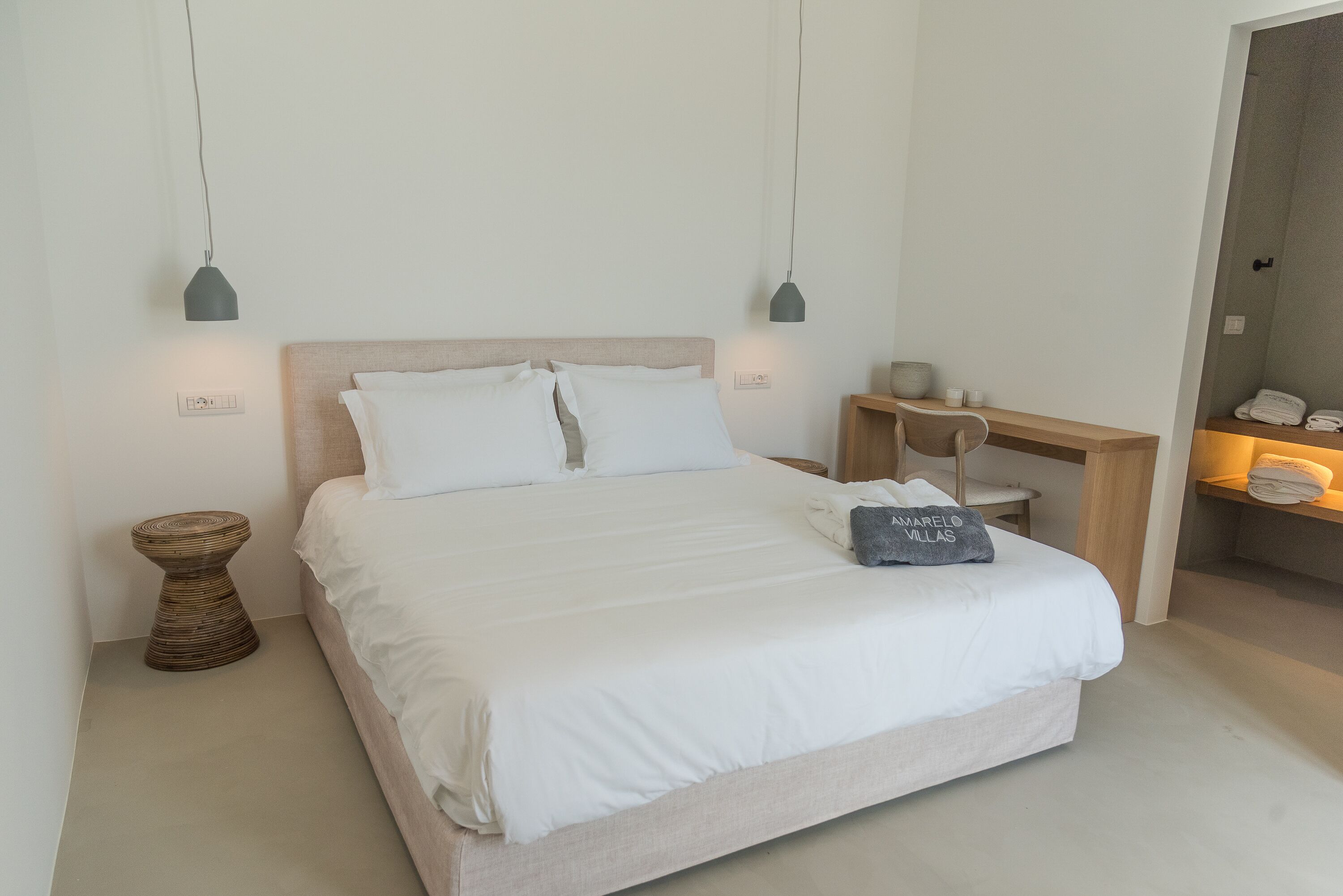 A neatly made bed with white linens and a pillow with text, a bedside table with a lamp, and a small round stool.