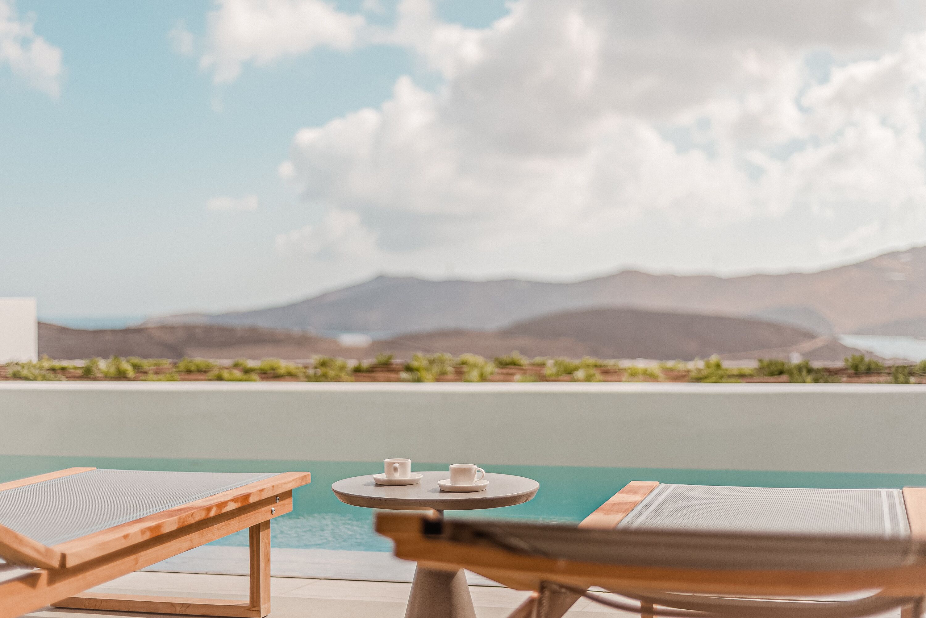 A poolside area with lounge chairs and a small table with cups, overlooking a body of water and distant mountains.