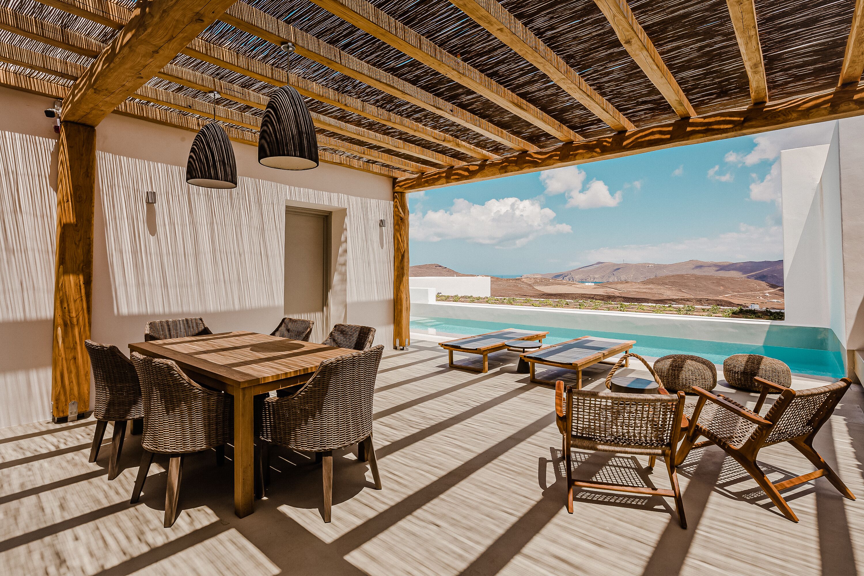 Un patio al aire libre con mesa de madera y sillas de mimbre, con vistas a una piscina y montañas.