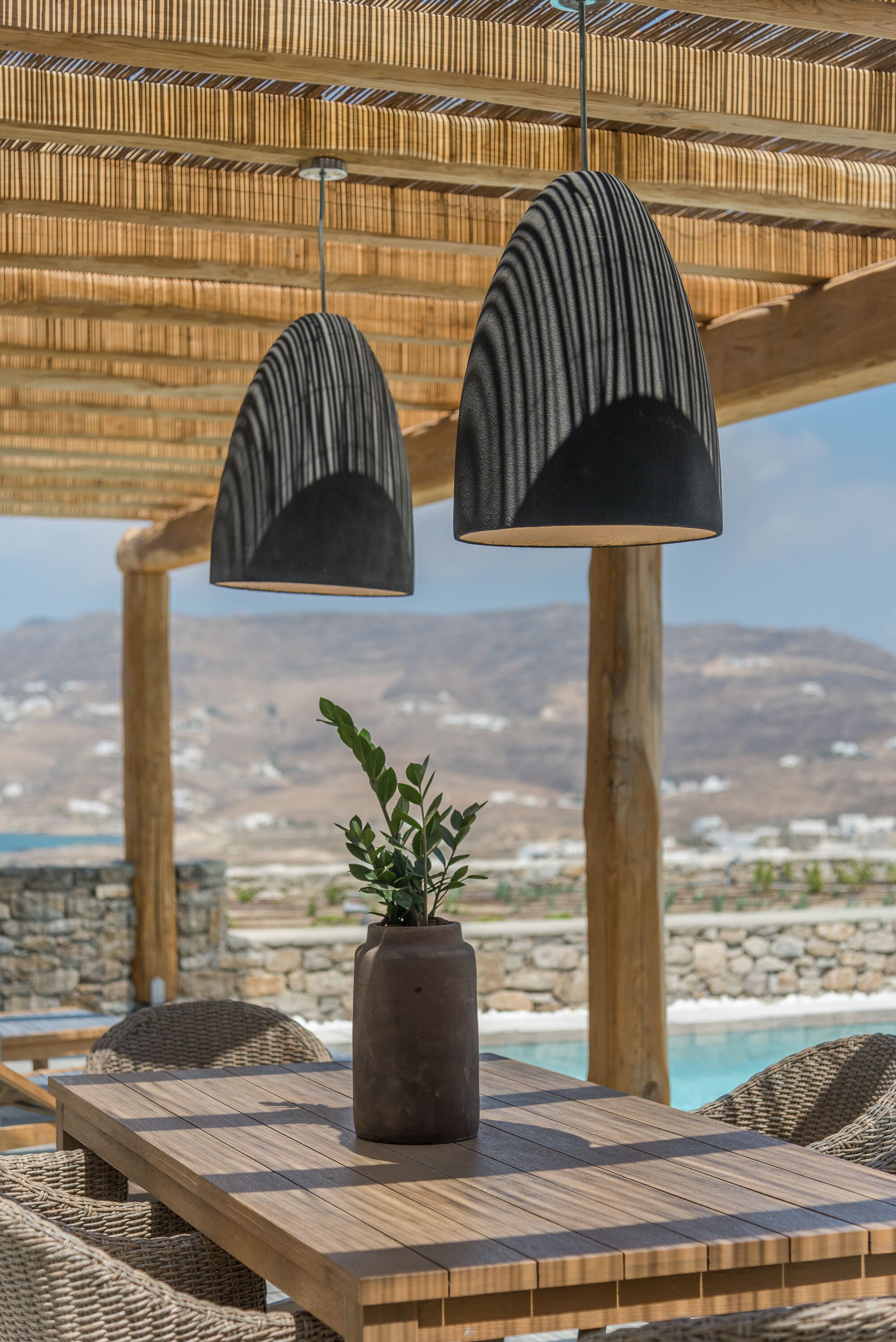 A wooden table with a potted plant, two hanging lamps, wicker chairs, and a view of a mountainous landscape.