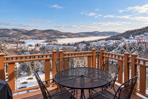 A balcony with a round table and chairs, overlooking a snowy landscape with buildings and a lake.