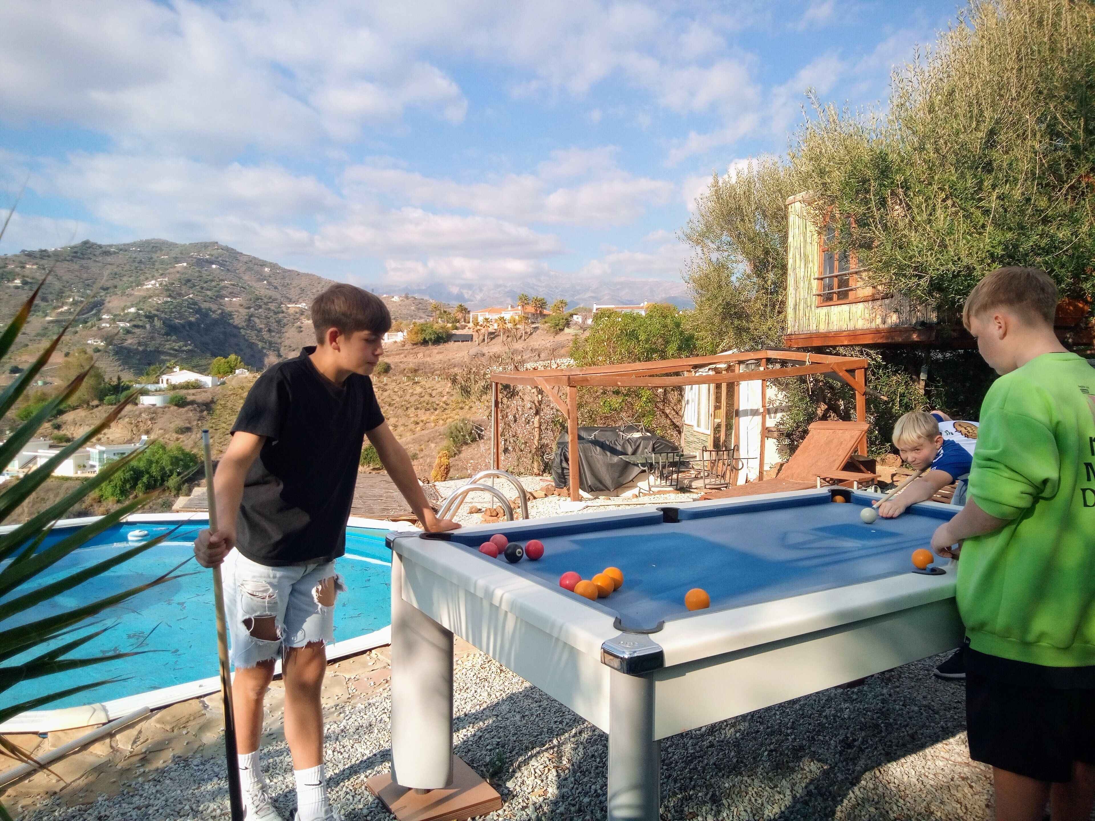 An outdoor pool area with a pool table, a pool, and a view of a hilly landscape.