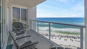 Varanda com duas cadeiras e uma mesa, com vista para uma praia com guarda-sóis e pessoas.