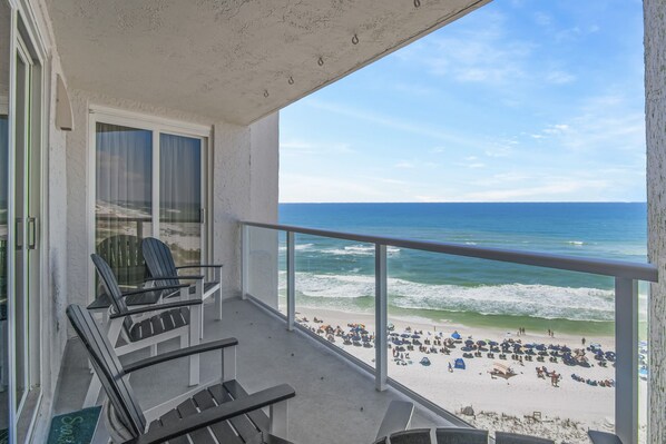 A balcony with two chairs and a table, overlooking a beach with umbrellas and people.
