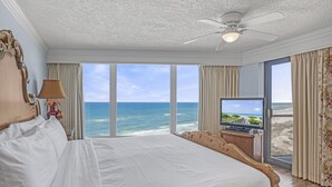 Uma cama grande com vista para o oceano, um ventilador de teto e uma televisão em uma mesinha de cabeceira de madeira.