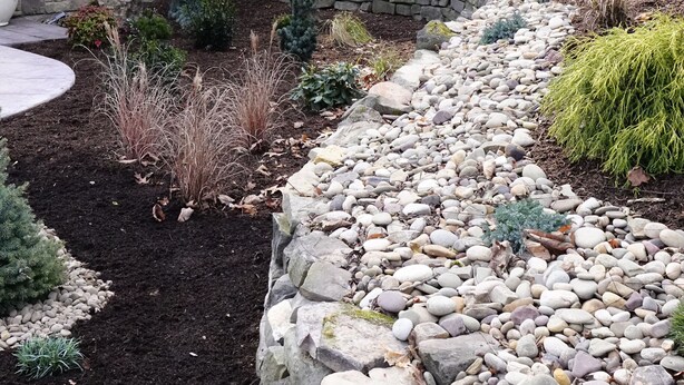 Een rustieke chalet met een stenen fundering en een rood dak, omgeven door een aangelegde tuin met een pad van stenen en diverse struiken.