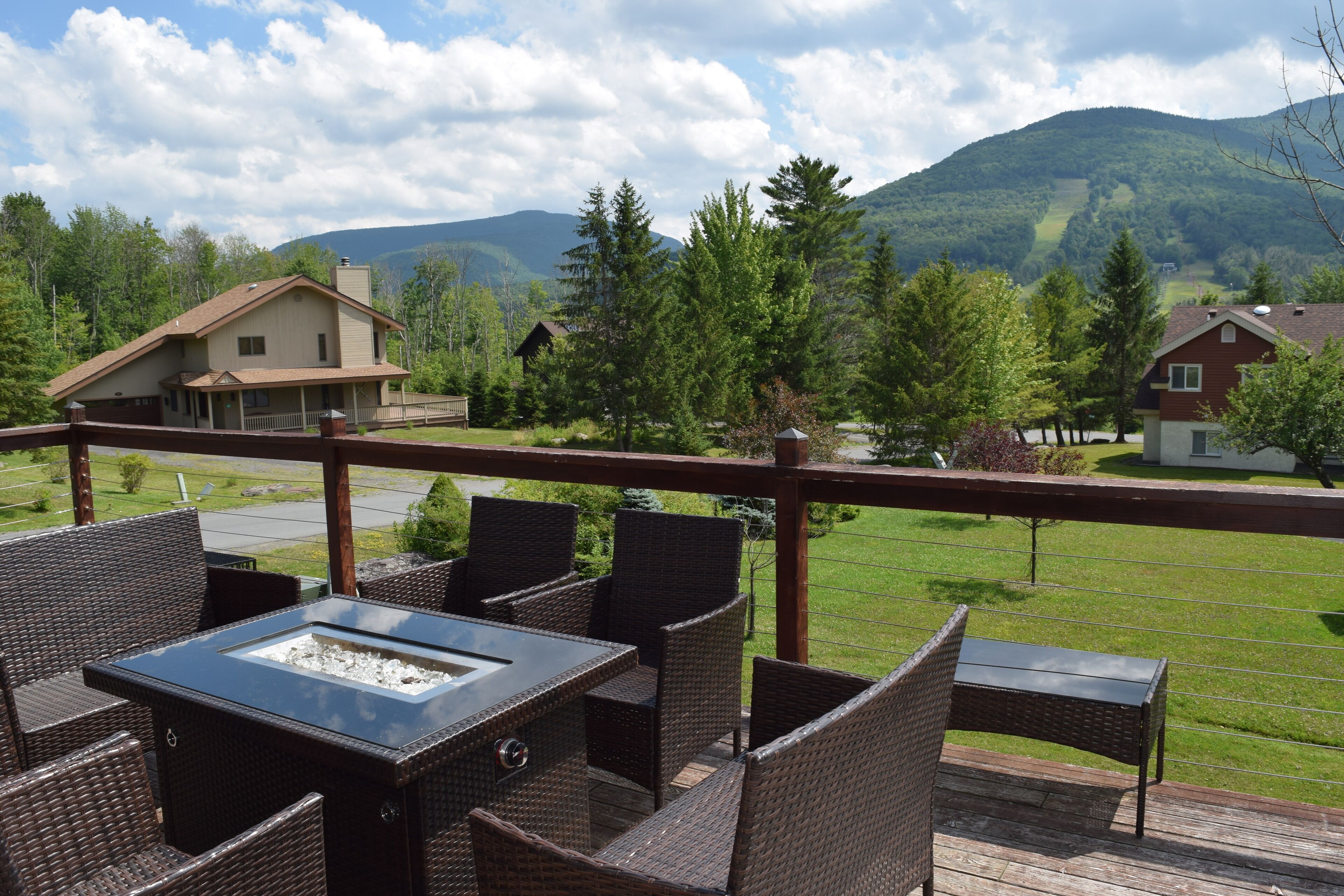 A deck with wicker furniture and a glass-top table, overlooking a green landscape with houses and mountains.