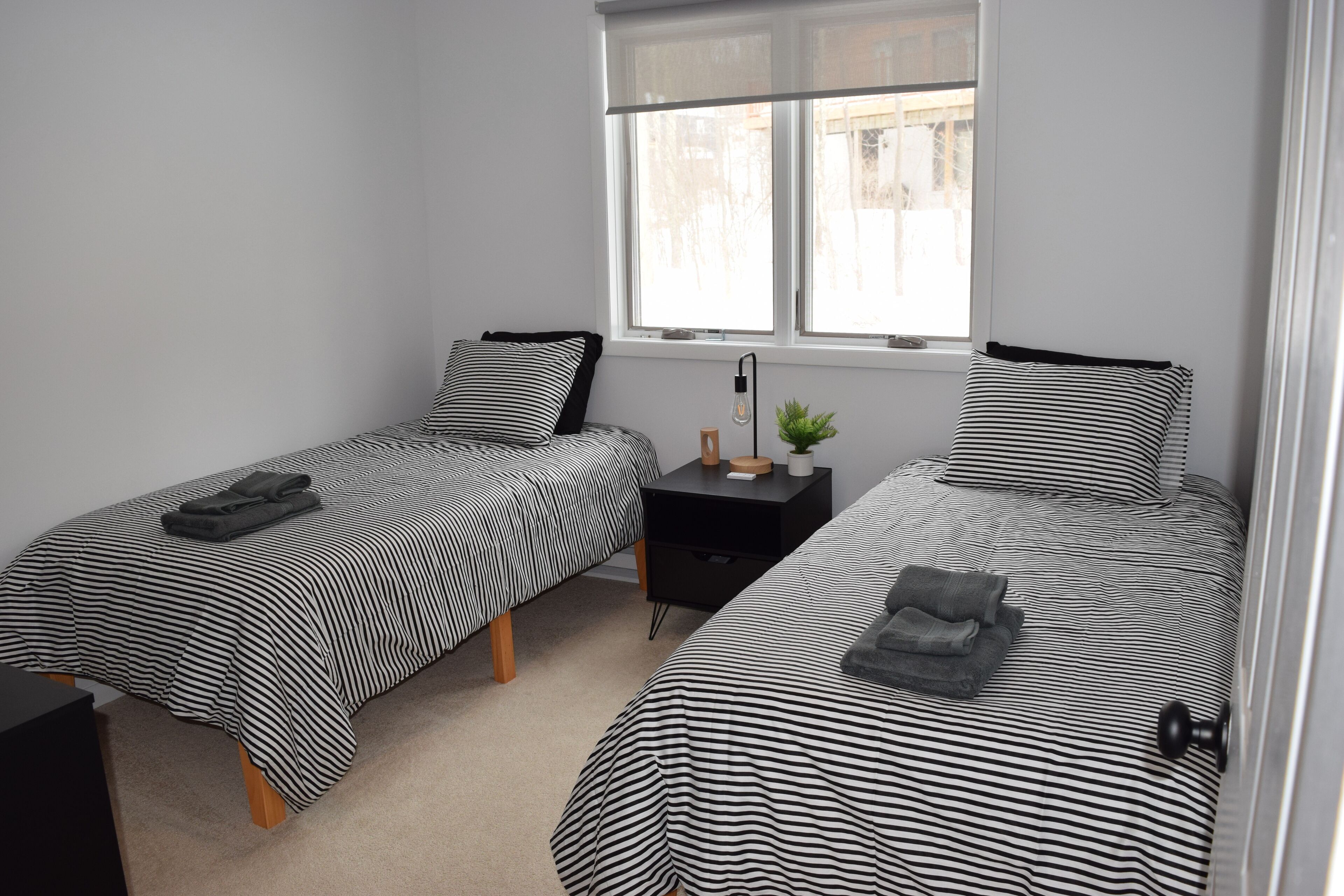 Une chambre double avec deux lits, chacun doté de draps à rayures, une petite table de chevet avec une lampe et une fenêtre donnant sur des immeubles.