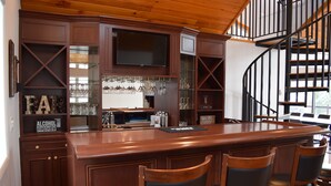 Uma área de bar com móveis de madeira, uma televisão presa acima do balcão e uma escadaria em espiral ao fundo.