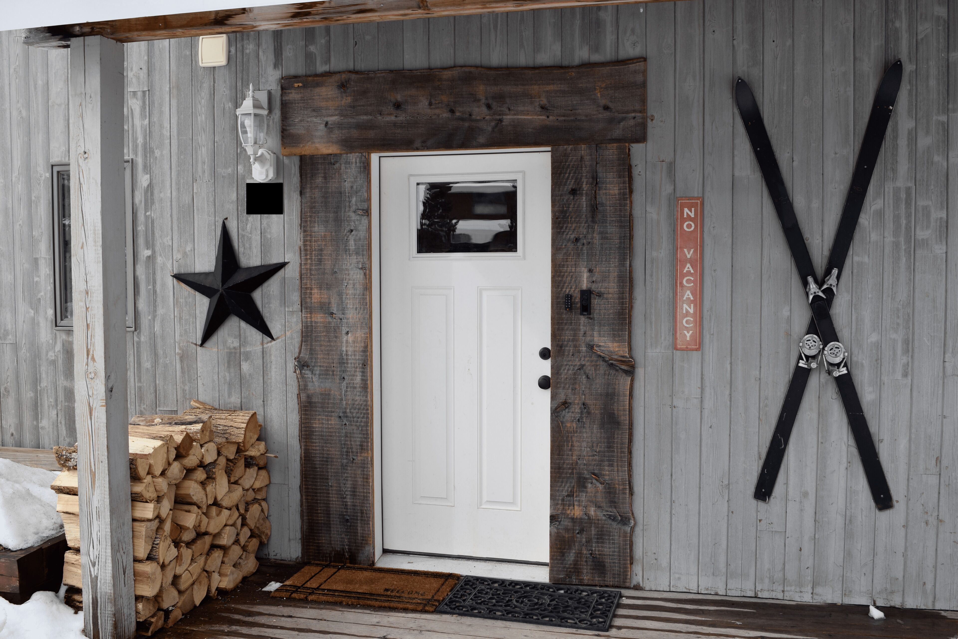 Un edificio rústico de madera con una puerta blanca, un adorno con forma de estrella y letreros que indican 'NO VACANCY', además de esquíes colgados en la pared.