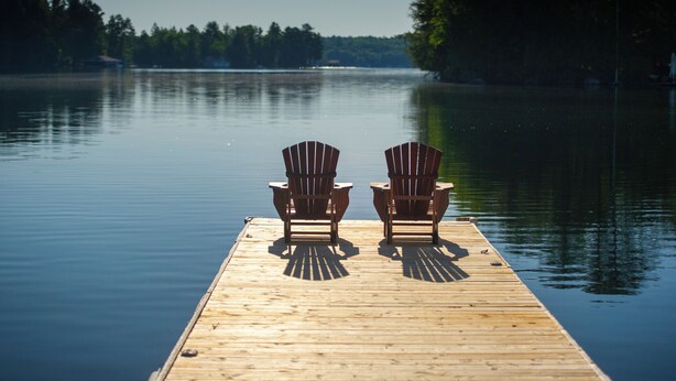 Um píer de madeira com duas cadeiras Adirondack voltadas para um lago calmo, cercado por árvores.