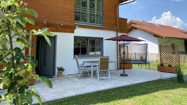 Een houten woonhuis met twee verdiepingen, met een terras, een tafel, stoelen, een trampoline en een tuin.