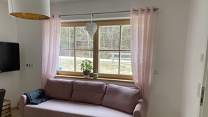 Een woonkamer met een bank, een aan de muur bevestigde televisie, een raam met gordijnen, een hanglamp en een kamerplant.