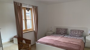 Een slaapkamer met een bed, een nachtkastje, een stoel, een raam met gordijnen en een hartdecoratie aan de muur.